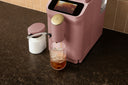 A pink countertop beverage dispenser with a digital display screen and a gold-toned dispensing head, shown dispensing an iced drink into a clear glass on a dark granite kitchen counter.