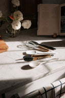 Set of kitchen utensils on a marble surface with flowers and an open book in the background.