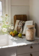 Kitchen counter with a jar, apples, and flowers near a window.