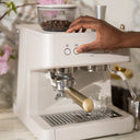 White espresso machine with a hand adjusting settings, blurred floral background